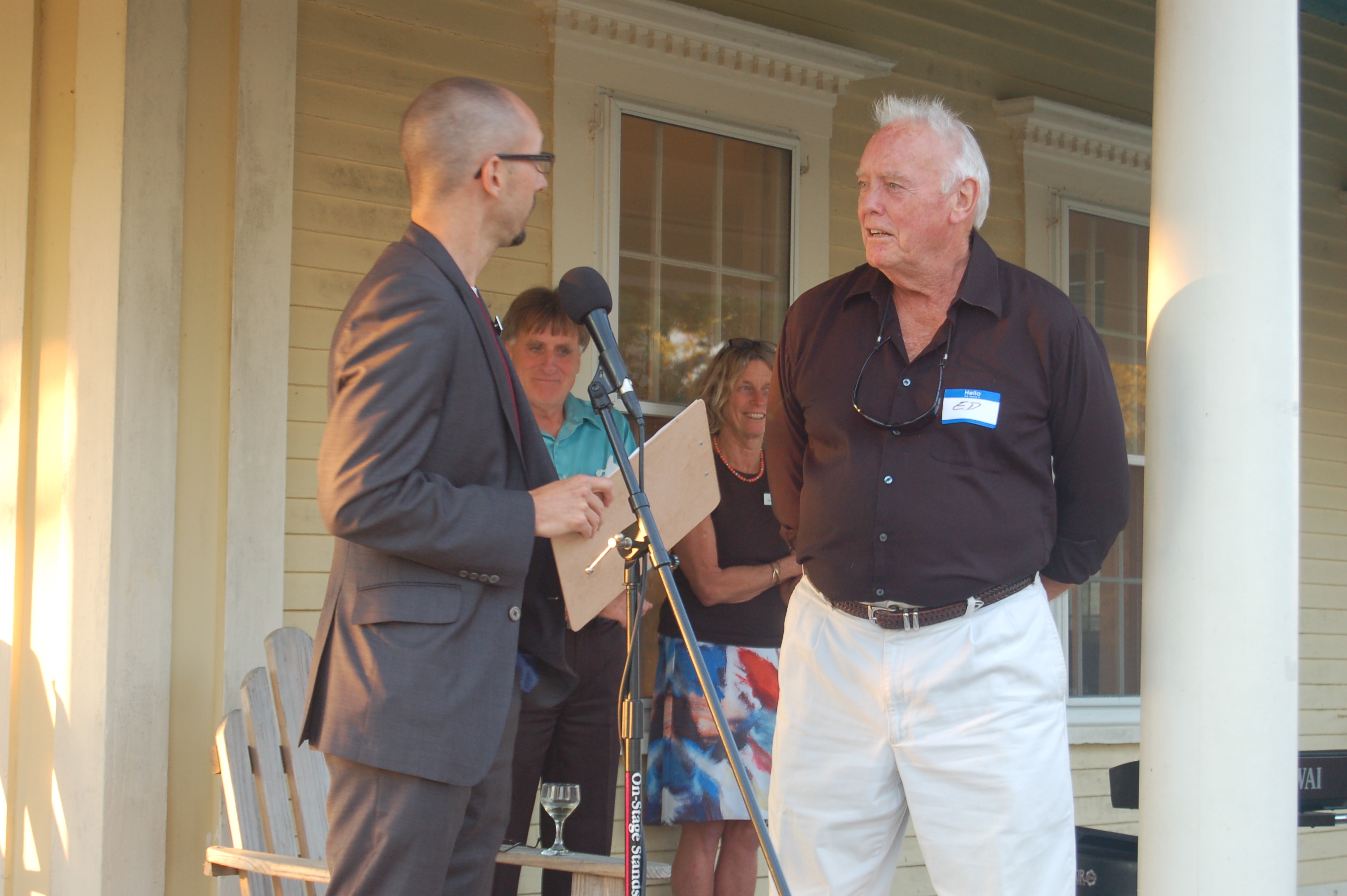 Ed Bellegarde (right) accepts a Volunteer Extraordinaire award from Nik Charov, president of Laudholm Trust, at the 2014 volunteer recognition event. [Two men in the foreground, a man and woman in the background, on a large porch.]