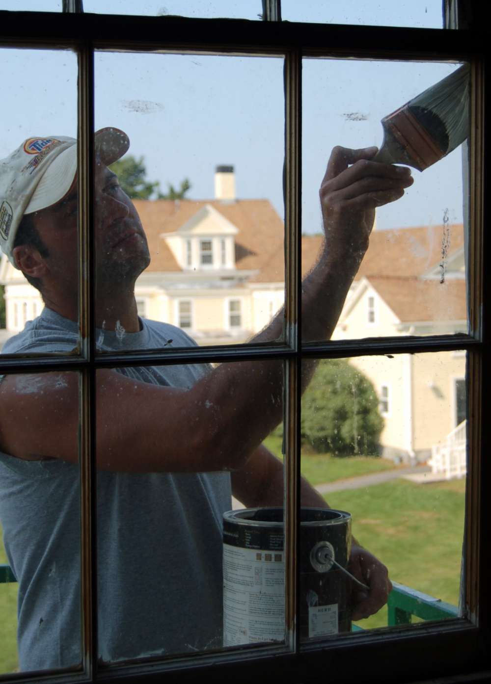 Through a window, a painter is seen applying a brush to the frame. The Laudholm farmhouse is in the background.